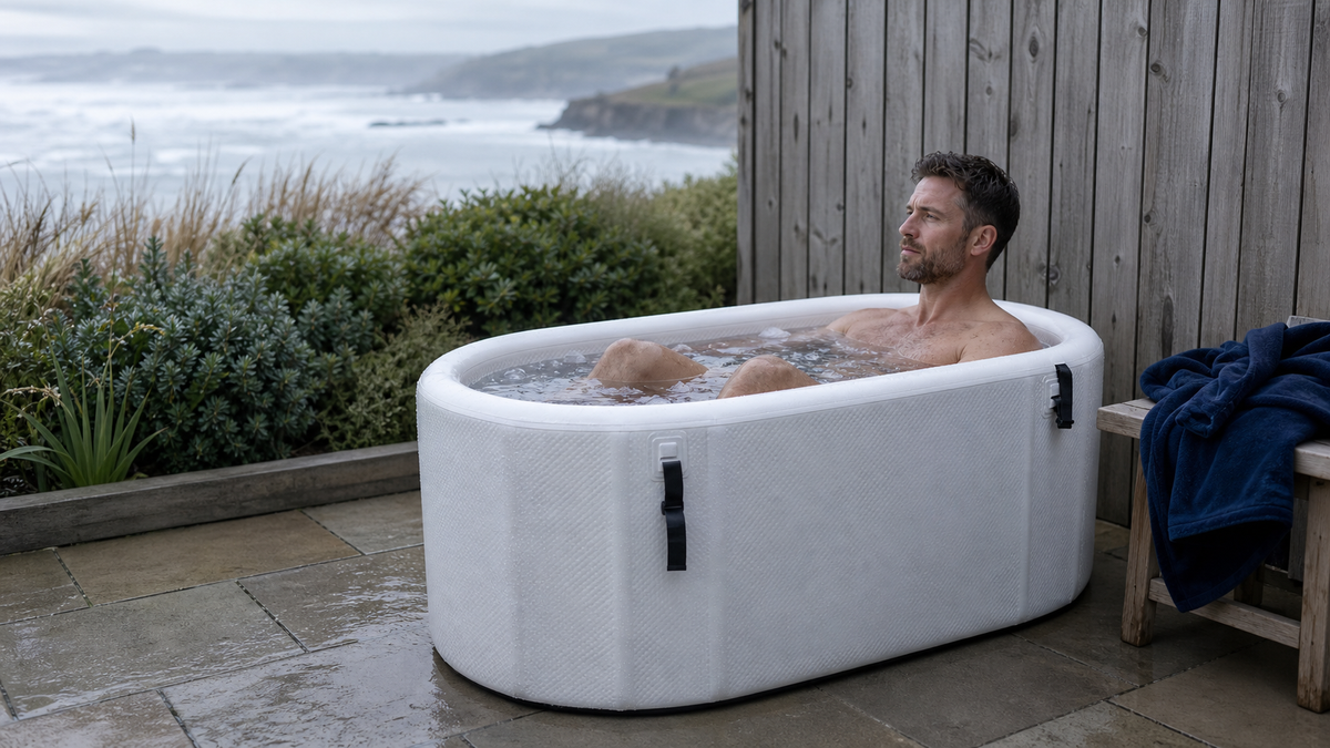 Man seated lengthways in a long white ice bath on a British coastal garden terrace